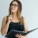 Mulher pensativa com óculos segura caderno e caneta.