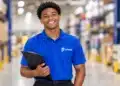 Jovem negro com camisa azul da Praso e prancheta, em um depósito.
