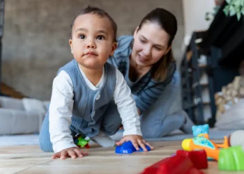 Bebê engatinha com brinquedos coloridos enquanto mulher sorri ao fundo.