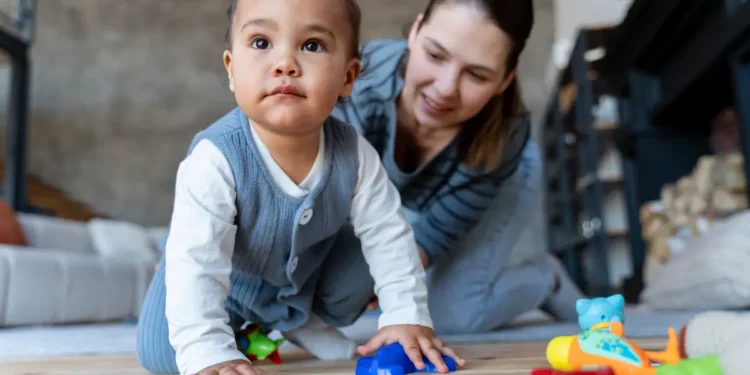 Bebê engatinha com brinquedos coloridos enquanto mulher sorri ao fundo.