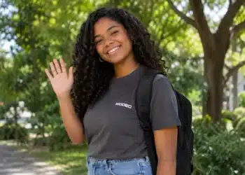 Jovem sorrindo e acenando, com camiseta da MODEC e mochila nas costas.