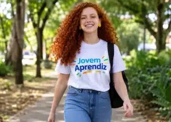 Jovem ruiva sorridente com camiseta “Jovem Aprendiz” e calça jeans, caminhando em meio às árvores.