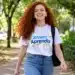 Jovem ruiva sorridente com camiseta “Jovem Aprendiz” e calça jeans, caminhando em meio às árvores.