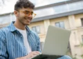 Jovem sorrindo enquanto usa um notebook ao ar livre, representando a inscrição online no Vestibular Cederj 2027.