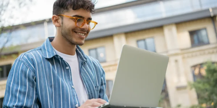 Jovem sorrindo enquanto usa um notebook ao ar livre, representando a inscrição online no Vestibular Cederj 2027.