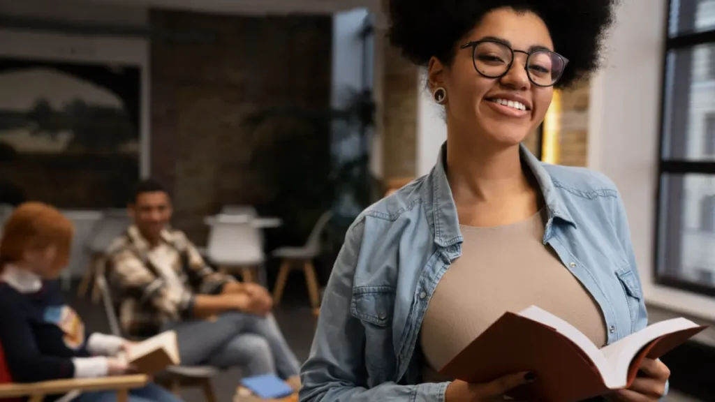 Jovem sorrindo com um livro nas mãos, com colegas estudando ao fundo, representando a preparação para o Vestibular Cederj 2027.