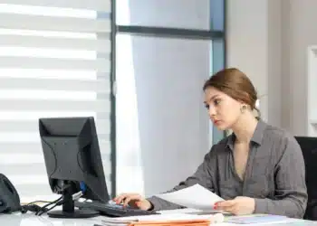 Jovem auxiliar administrativo trabalha no computador com documentos sobre a mesa do escritório