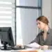 Jovem auxiliar administrativo trabalha no computador com documentos sobre a mesa do escritório