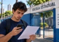 Estudante mexe no celular e segura um papel em frente a uma escola, representando a consulta ao local de prova da reaplicação do Encceja 2026.