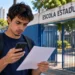 Estudante mexe no celular e segura um papel em frente a uma escola, representando a consulta ao local de prova da reaplicação do Encceja 2026.