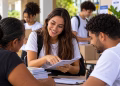 Imagem ilustrativa de universitária sorridente mostrando um folheto a moradores em uma mesa de atendimento, com outros estudantes de camiseta branca ao fundo, em referência à curricularização da extensão