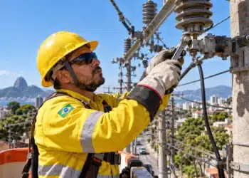 Eletricista com capacete amarelo, óculos de proteção e luvas de segurança realiza manutenção em equipamentos de uma rede elétrica instalada em poste.