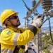 Eletricista com capacete amarelo, óculos de proteção e luvas de segurança realiza manutenção em equipamentos de uma rede elétrica instalada em poste.