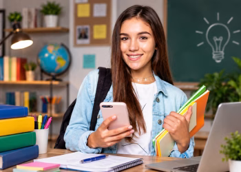 Imagem ilustrativa de jovem sorridente com celular em uma das mãos e pastas coloridas na outra, diante de uma mesa com livros, caderno e notebook, em referência aos cursos gratuitos para o Enem 2026