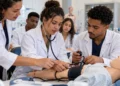 Grupo de quatro estudantes em jaleco usa estetoscópio e esfigmomanômetro em manequim humano em laboratório, simbolizando as vagas em cursos técnicos na área da saúde.