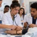 Grupo de quatro estudantes em jaleco usa estetoscópio e esfigmomanômetro em manequim humano em laboratório, simbolizando as vagas em cursos técnicos na área da saúde.