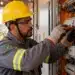 Eletricista brasileiro com capacete amarelo e equipamentos de proteção realiza manutenção em quadro de distribuição de energia elétrica.
