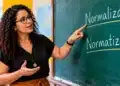 Professora aponta para as palavras normalizar e normatizar escritas no quadro-negro durante explicação em sala de aula.