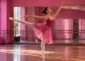 Bailarina com vestido rosa executa movimento de balé na ponta dos pés em estúdio de dança com espelhos.
