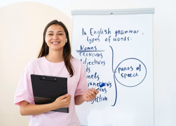 Imagem ilustrativa de professora sorrindo ao lado de um quadro com anotações sobre gramática inglesa, segurando uma prancheta, em referência à formação gratuita para professores de inglês
