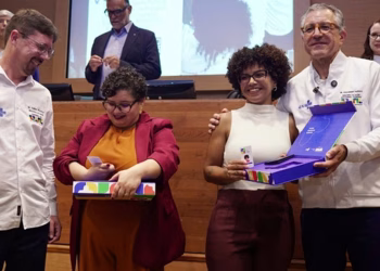 Profissionais sorridentes seguram cartões e uma caixa azul em cerimônia, ao lado de homens com camisas do SUS, em referência à emissão das carteiras da profissão de sanitarista