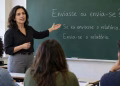 Professora gesticula para lousa com "Enviasse ou envia-se?" ao ensinar alunos em classe, explicando a dúvida gramatical.