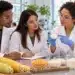 Três cientistas com jalecos e óculos analisam amostra em laboratório com milho e polvilho sobre mesa, simbolizando o estágio Ingredion.