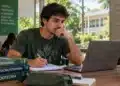 Estudante de Agronomia sentado em ambiente universitário, olhando para o notebook e segurando uma caneta sobre o caderno.