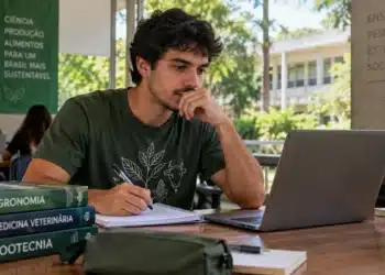 Estudante de Agronomia sentado em ambiente universitário, olhando para o notebook e segurando uma caneta sobre o caderno.