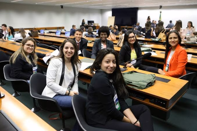 Jovens sorridentes sentados em bancada de plenário de comissão da Câmara dos Deputados, com microfones e mesas de madeira, em referência ao estágio na Câmara dos Deputados