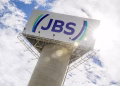 Logo da JBS em placa no alto de uma torre de concreto, com céu azul e sol ao fundo, ilustrando a matéria sobre o estágio na JBS