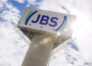 Logo da JBS em placa no alto de uma torre de concreto, com céu azul e sol ao fundo, ilustrando a matéria sobre o estágio na JBS
