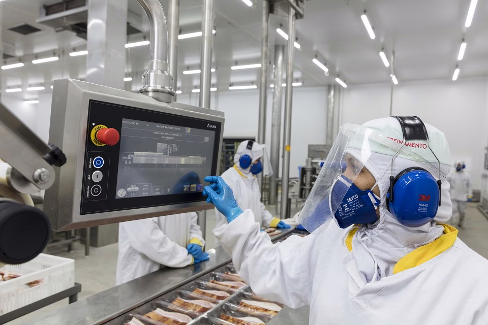 Imagem ilustrativa de funcionária com uniforme branco, protetor facial e abafador de ruído operando painel digital em linha de produção, ligada às vagas de estágio na JBS