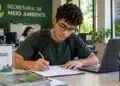 Jovem estudante preenchendo formulário em uma mesa de escritório, com notebook e identificação de estágio na Secretaria de Meio Ambiente.