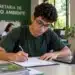 Jovem estudante preenchendo formulário em uma mesa de escritório, com notebook e identificação de estágio na Secretaria de Meio Ambiente.