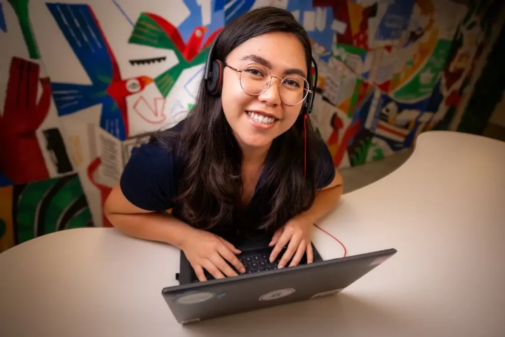 Jovem de óculos e fones de ouvido utiliza um notebook sobre uma mesa, com um mural colorido ao fundo.