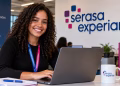 Imagem ilustrativa de jovem de cabelos cacheados com crachá, sorrindo diante de um notebook em escritório com o logo da Serasa Experian na parede, representando o estágio Serasa Experian