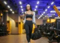 Mulher com roupa esportiva olha para o celular enquanto caminha em uma academia, carregando uma bolsa.
