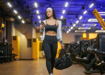 Mulher com roupa esportiva olha para o celular enquanto caminha em uma academia, carregando uma bolsa.