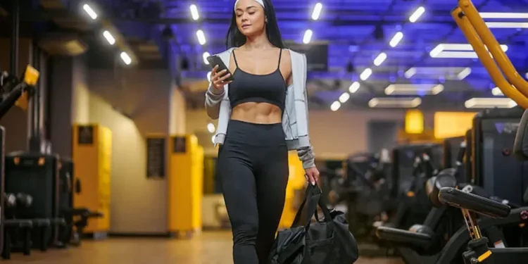 Mulher com roupa esportiva olha para o celular enquanto caminha em uma academia, carregando uma bolsa.