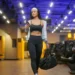Mulher com roupa esportiva olha para o celular enquanto caminha em uma academia, carregando uma bolsa.