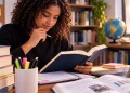 Imagem ilustrativa de estudante de cabelos cacheados lendo um livro de capa azul em uma mesa com cadernos, marca-textos, notebook e livros empilhados, em referência ao hábito de ler na preparação para o Enem