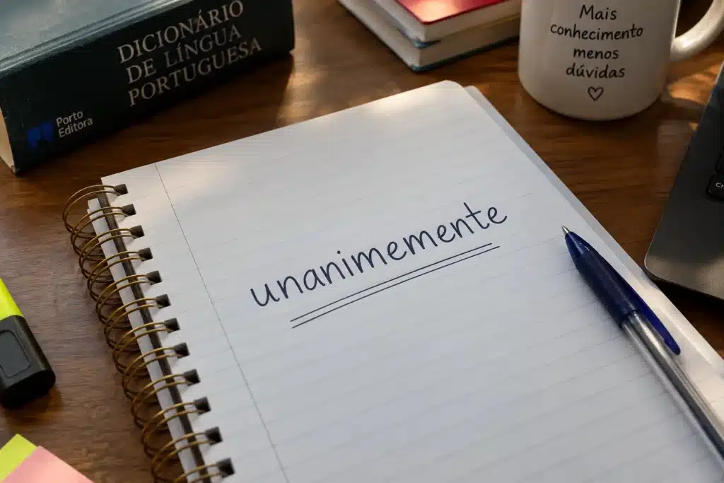 Caderno aberto sobre uma mesa de madeira com a palavra “unanimemente” escrita à mão e sublinhada, ao lado de uma caneta azul e um dicionário.