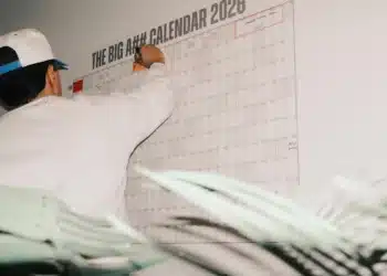 Homem anota datas em um grande calendário de 2026 na parede, planejando os feriados de novembro