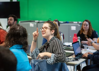 Pessoas em uma sala com notebooks, enquanto uma participante de óculos levanta o dedo para falar, em referência às vagas gratuitas do bootcamp do Itaú