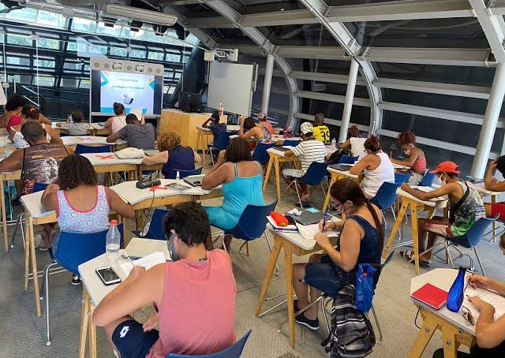 Alunos sentados em mesas fazem anotações durante aula em sala de vidro e estrutura metálica curva das Naves do Conhecimento