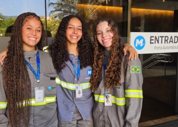 Três jovens aprendizes do MetrôRio, de uniforme cinza com faixas refletivas e crachá, sorriem abraçadas na entrada de uma unidade da empresa, participantes do Programa Jovem Aprendiz do MetrôRio