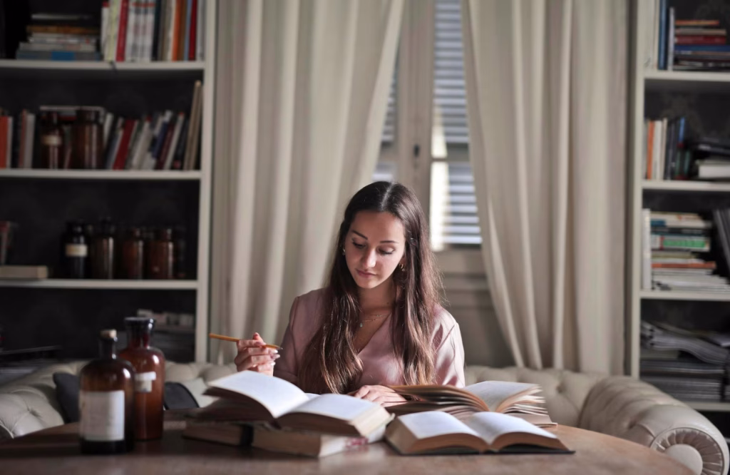 Imagem ilustrativa de estudante com lápis na mão lendo livros abertos sobre uma mesa, em frente a estantes, durante a preparação para o Vestibular Unicamp 2027