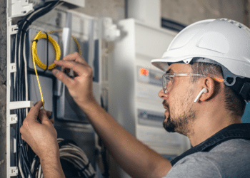 Imagem ilustrativa de eletricista de capacete branco e óculos de proteção organizando cabos num quadro elétrico, representando a manutenção elétrica predial