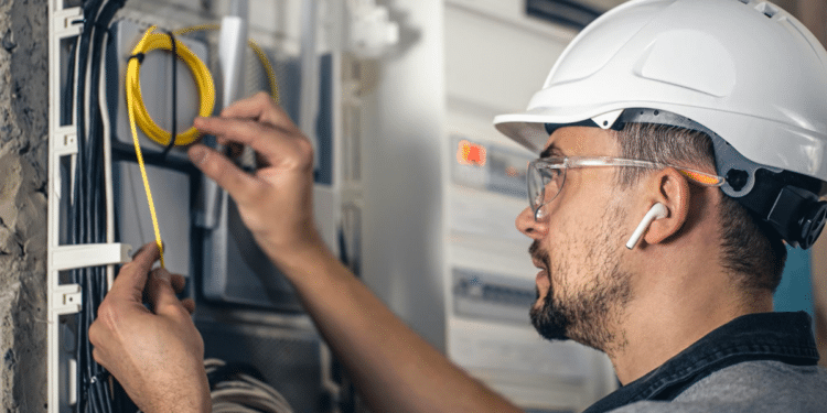 Imagem ilustrativa de eletricista de capacete branco e óculos de proteção organizando cabos num quadro elétrico, representando a manutenção elétrica predial